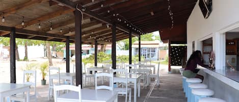Un café al aire libre con mesas y sillas blancas, una zona de bar con taburetes y un techo cubierto con luces colgantes.