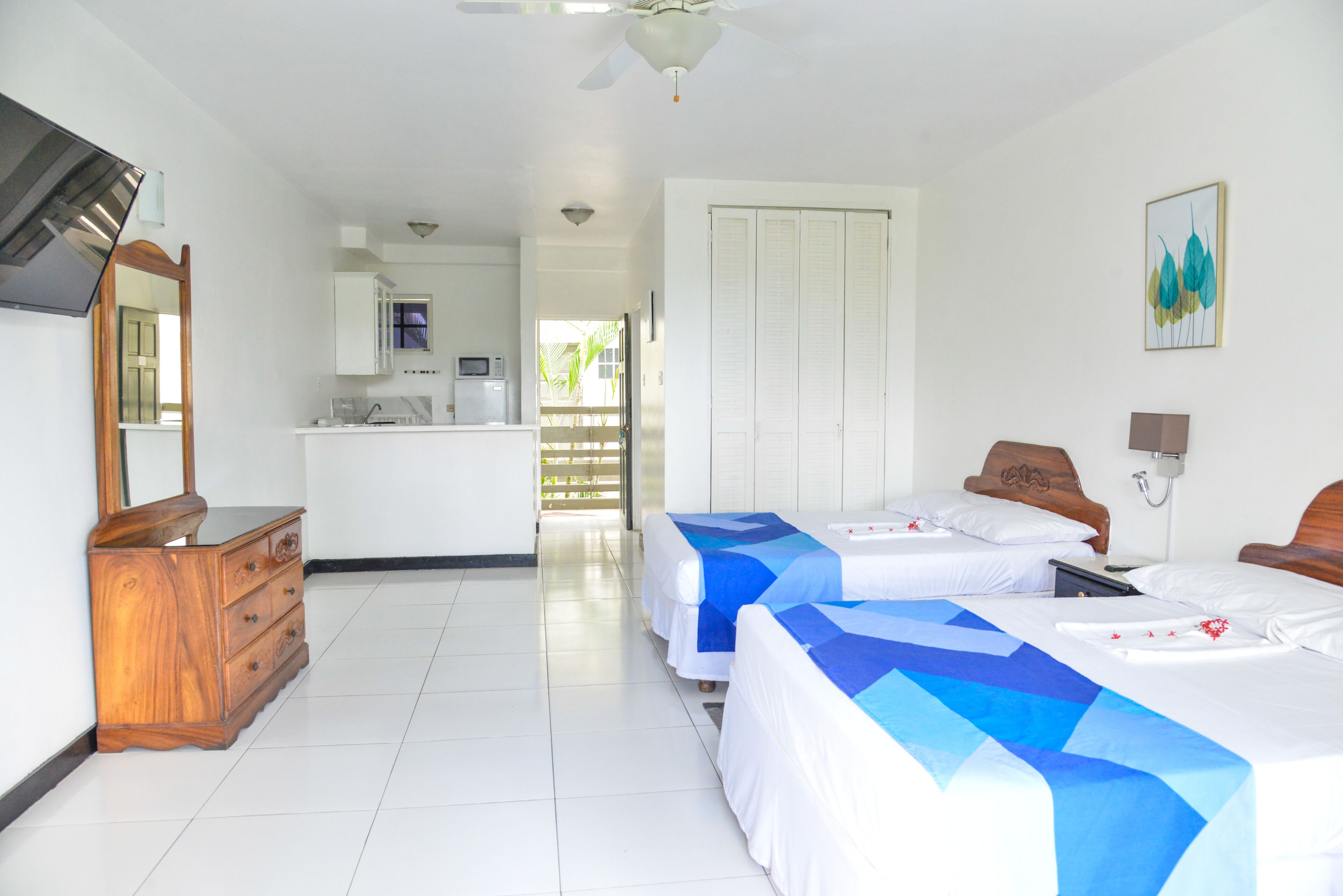 A hotel room with two beds, a wooden dresser, a kitchenette, and a door leading to a balcony.