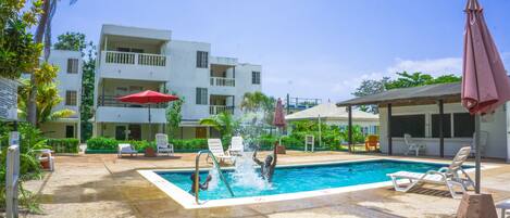 A swimming pool with lounge chairs and umbrellas, surrounded by a building with balconies and a covered area.