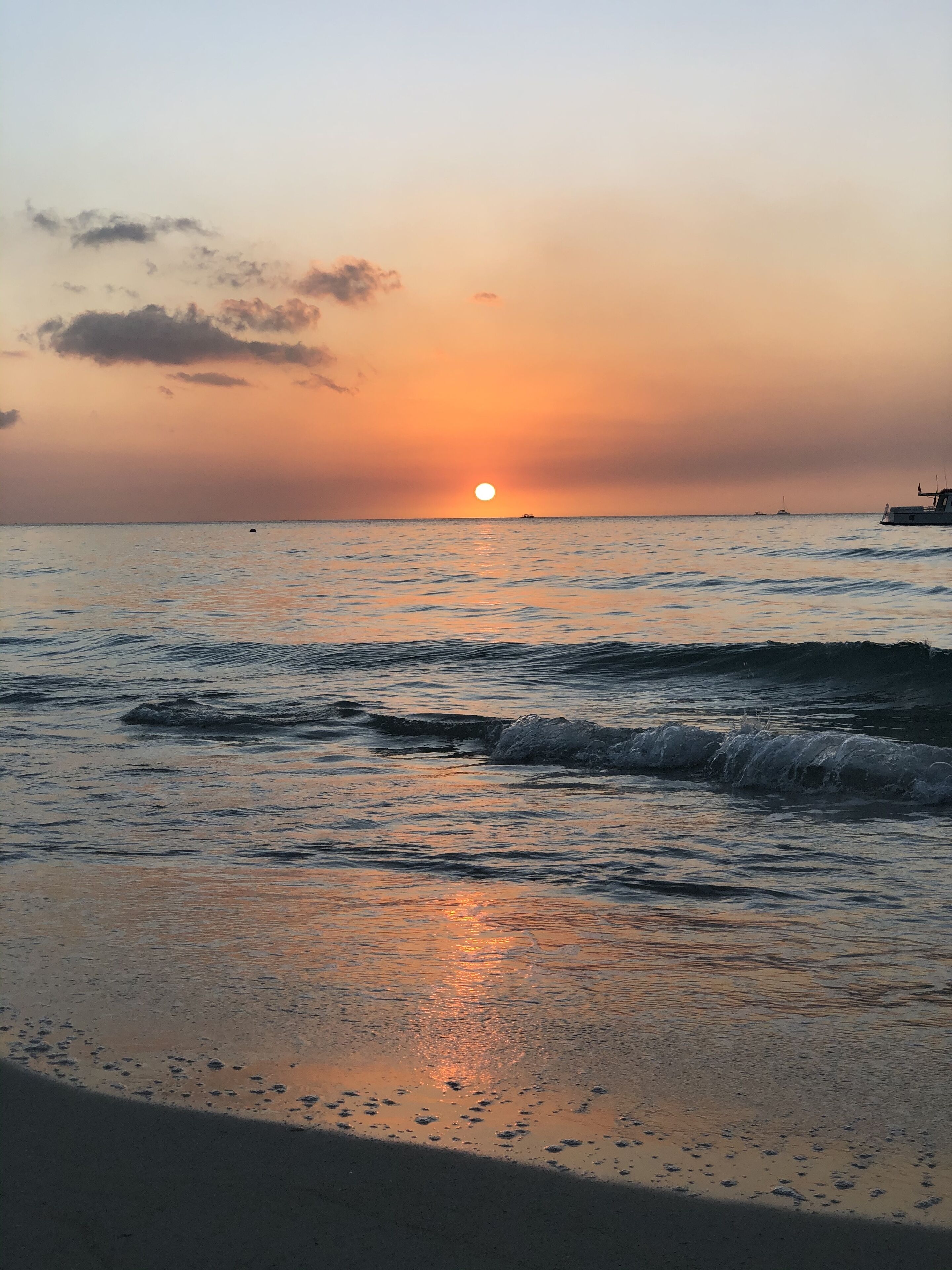 A beach scene with the sun setting over the ocean, waves gently lapping at the shore, and a boat in the distance.