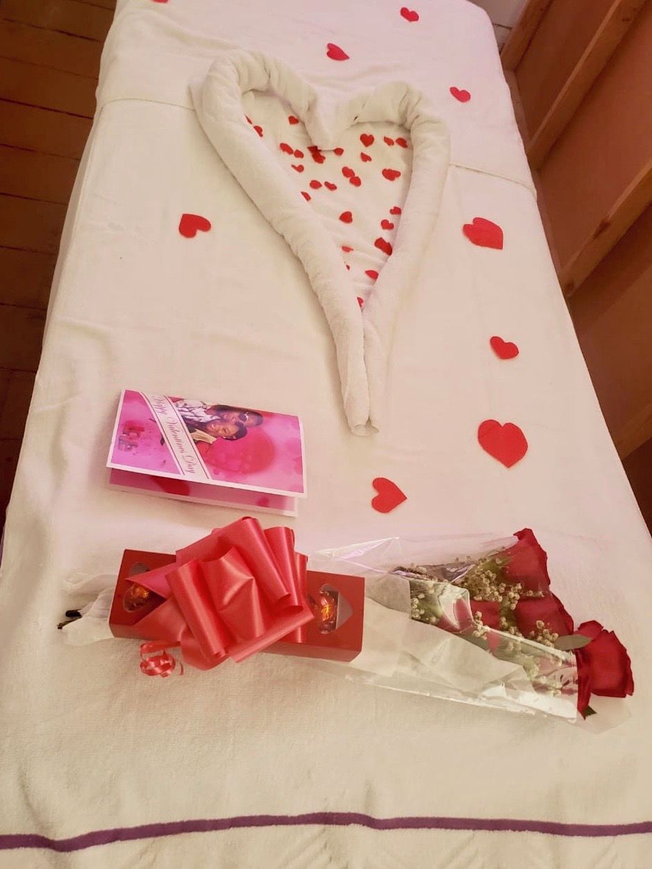 A bed with a heart-shaped pillow, a bouquet of roses, and a gift box.