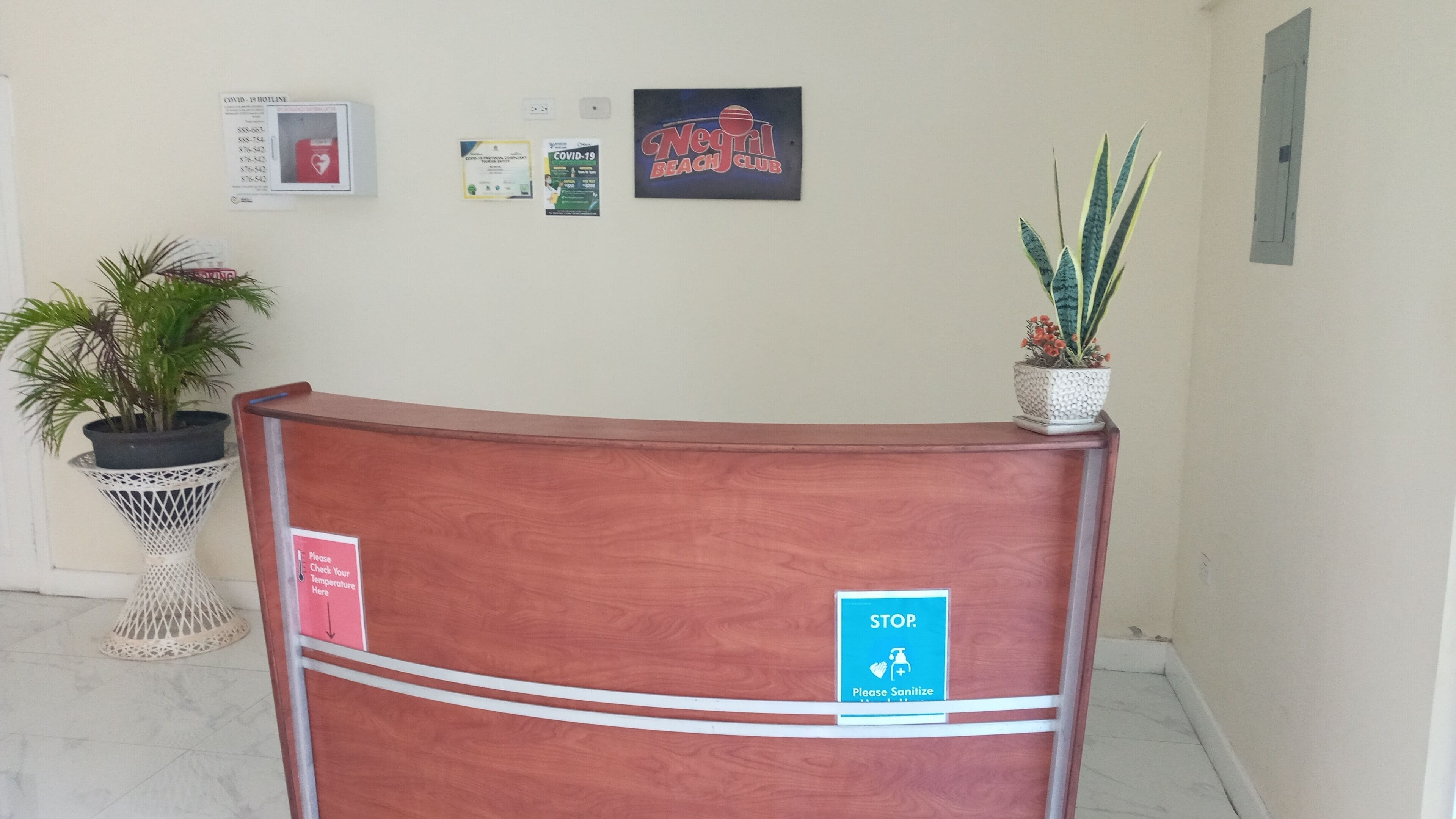 Un comptoir d’accueil avec un panneau indiquant « ARRÊT » et une affiche demandant aux visiteurs de se désinfecter les mains. Des plantes en pot et des affiches d’information sont disposées sur le mur.