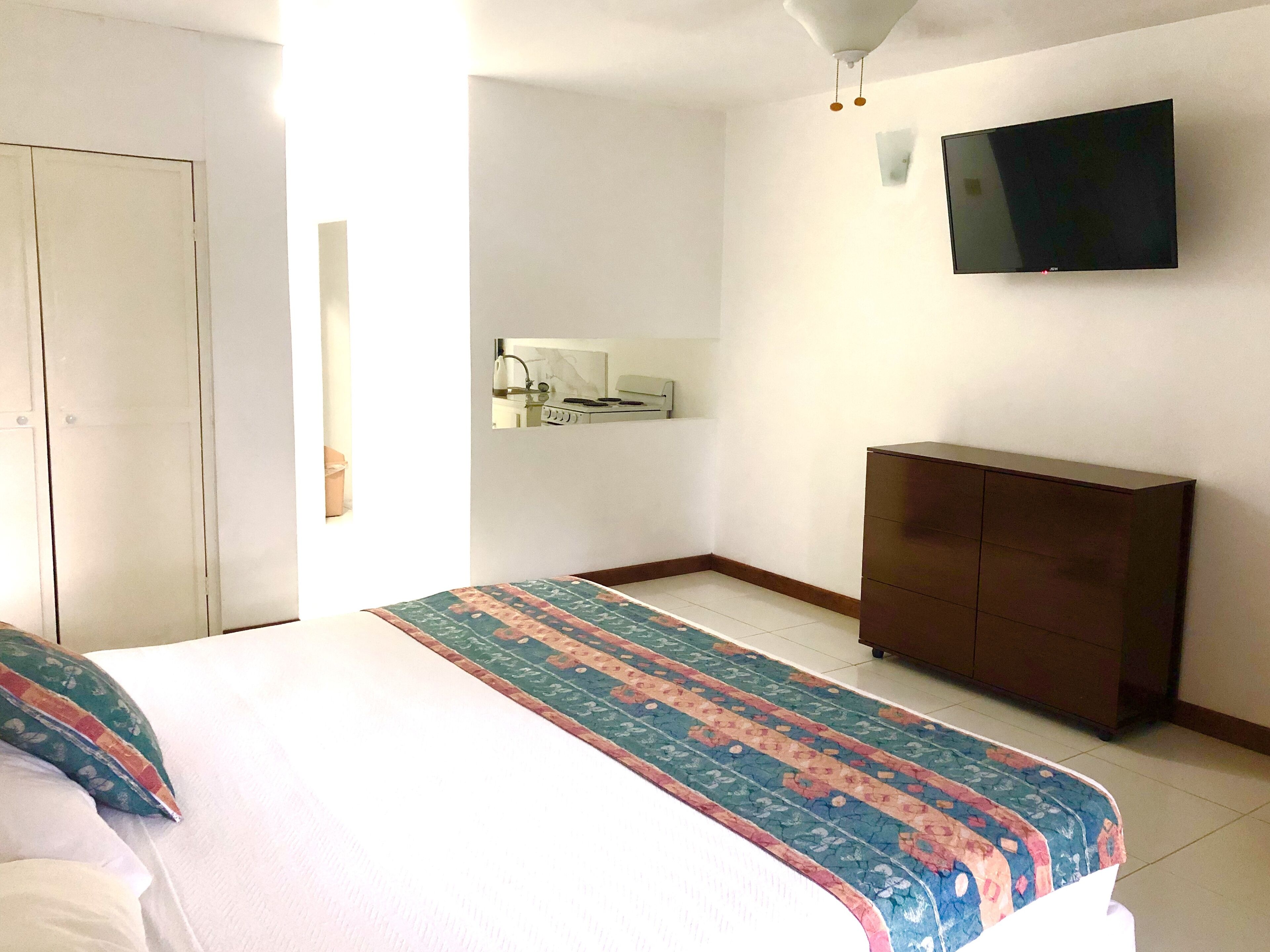 A bedroom with a bed, a dresser, a TV mounted on the wall, and a small kitchenette area.