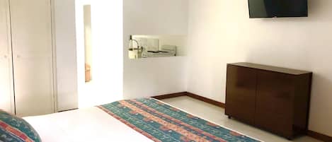 A bedroom with a bed, a dresser, a TV mounted on the wall, and a small kitchenette area.