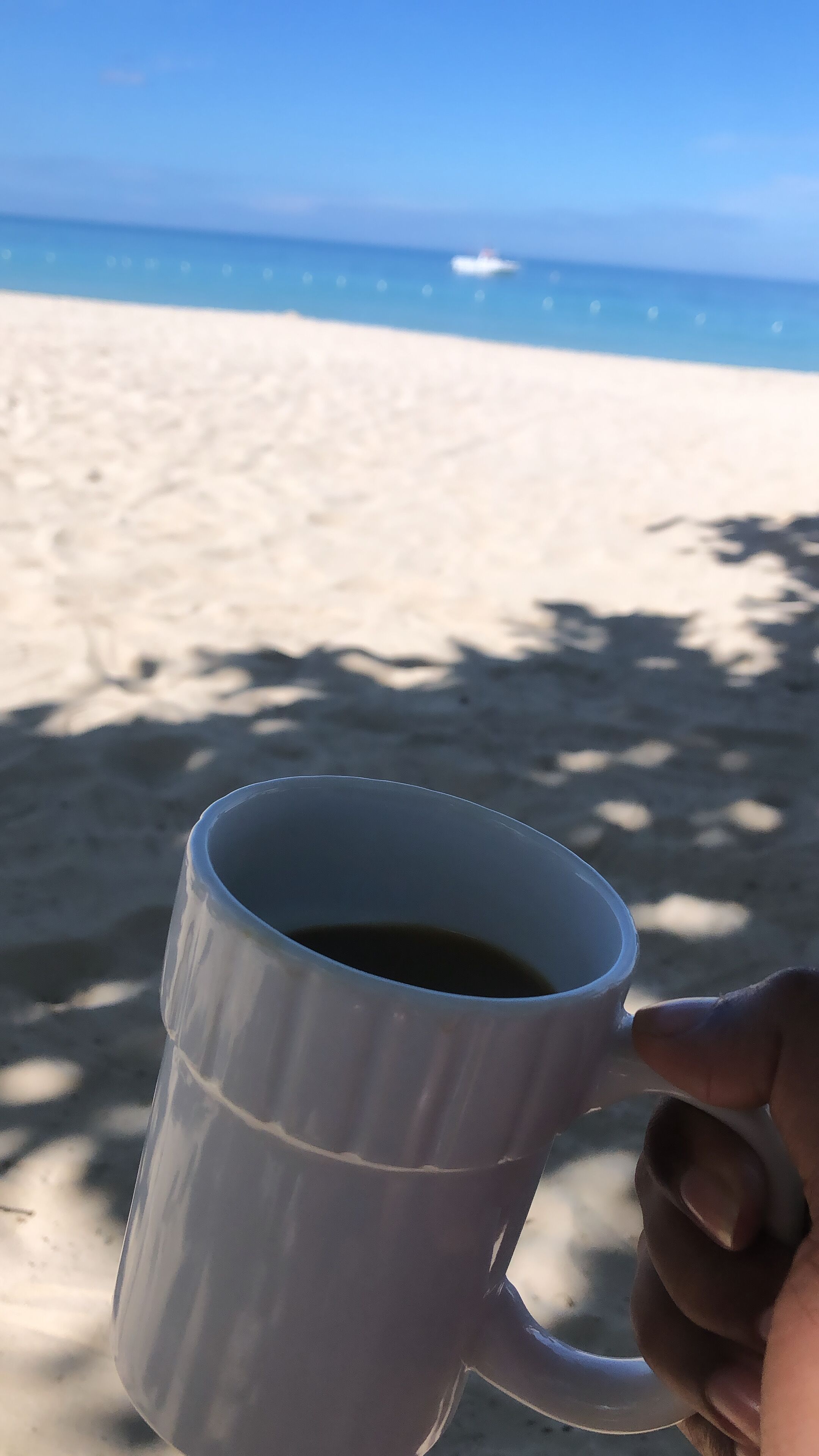 Una taza blanca con asa, sostenida por una mano, sobre una superficie arenosa con un cielo azul despejado y un barco distante al fondo.