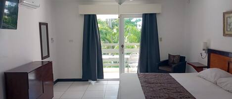 A hotel room with a bed, a desk, a chair, a ceiling fan, and a view of greenery outside.