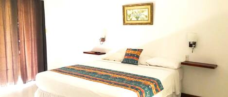 A hotel room with a bed, a nightstand, a wall-mounted lamp, a ceiling fan, and a window with curtains.