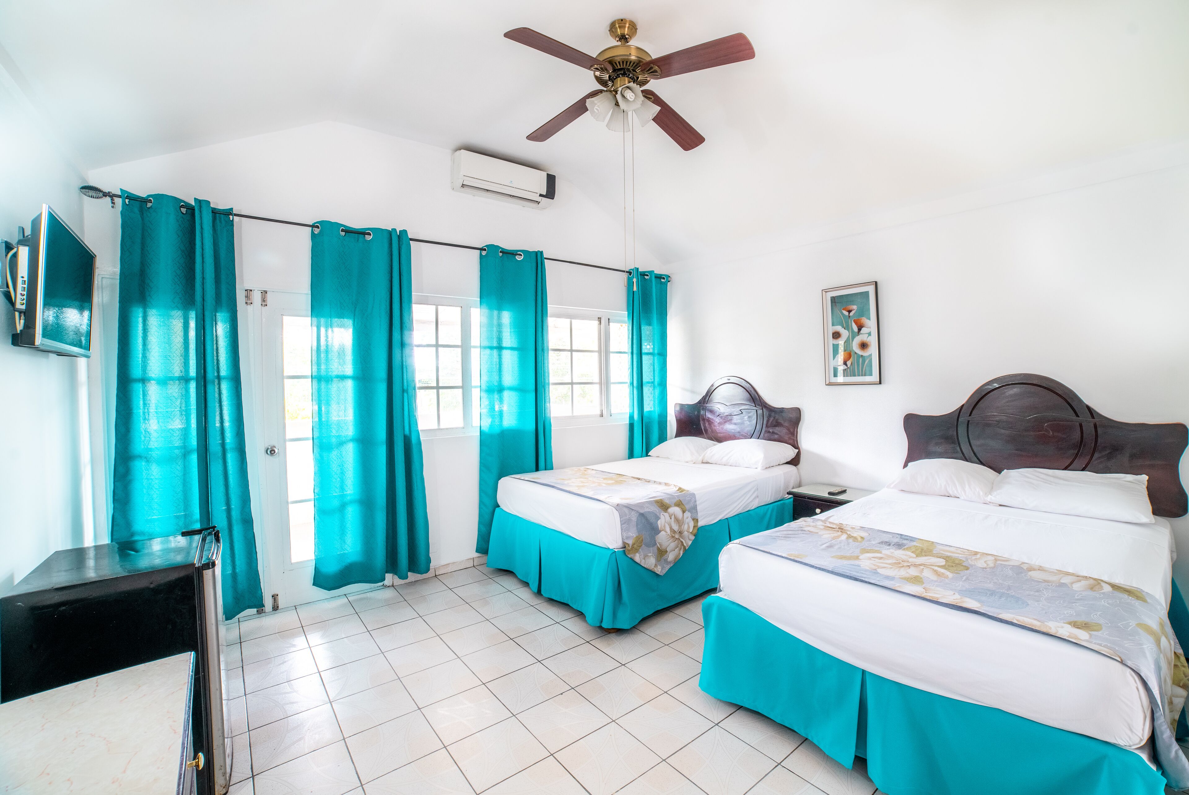 A hotel room with two beds, teal curtains, a ceiling fan, and a TV on the left wall.