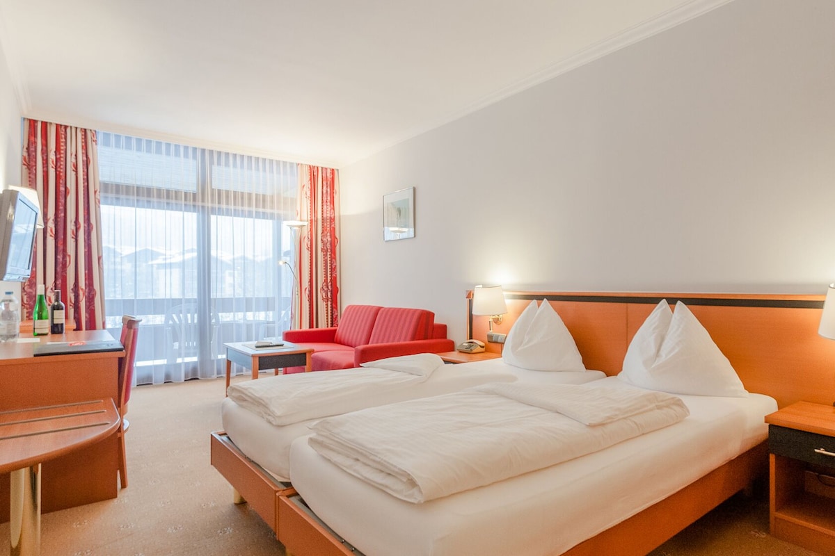 Quarto de hotel com cama, escrivaninha, cadeira, sofá vermelho e janela com cortinas.