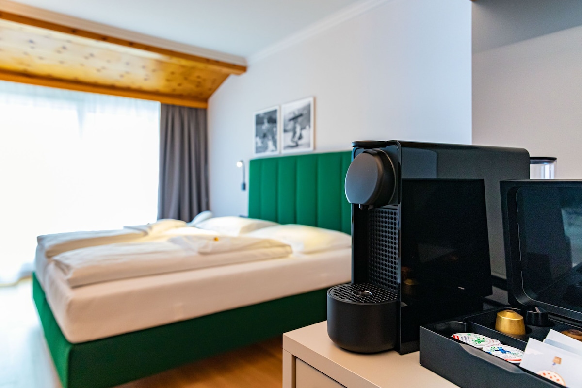 Quarto de hotel com cama, máquina de café, televisão e escrivaninha com uma xícara e alguns papéis.