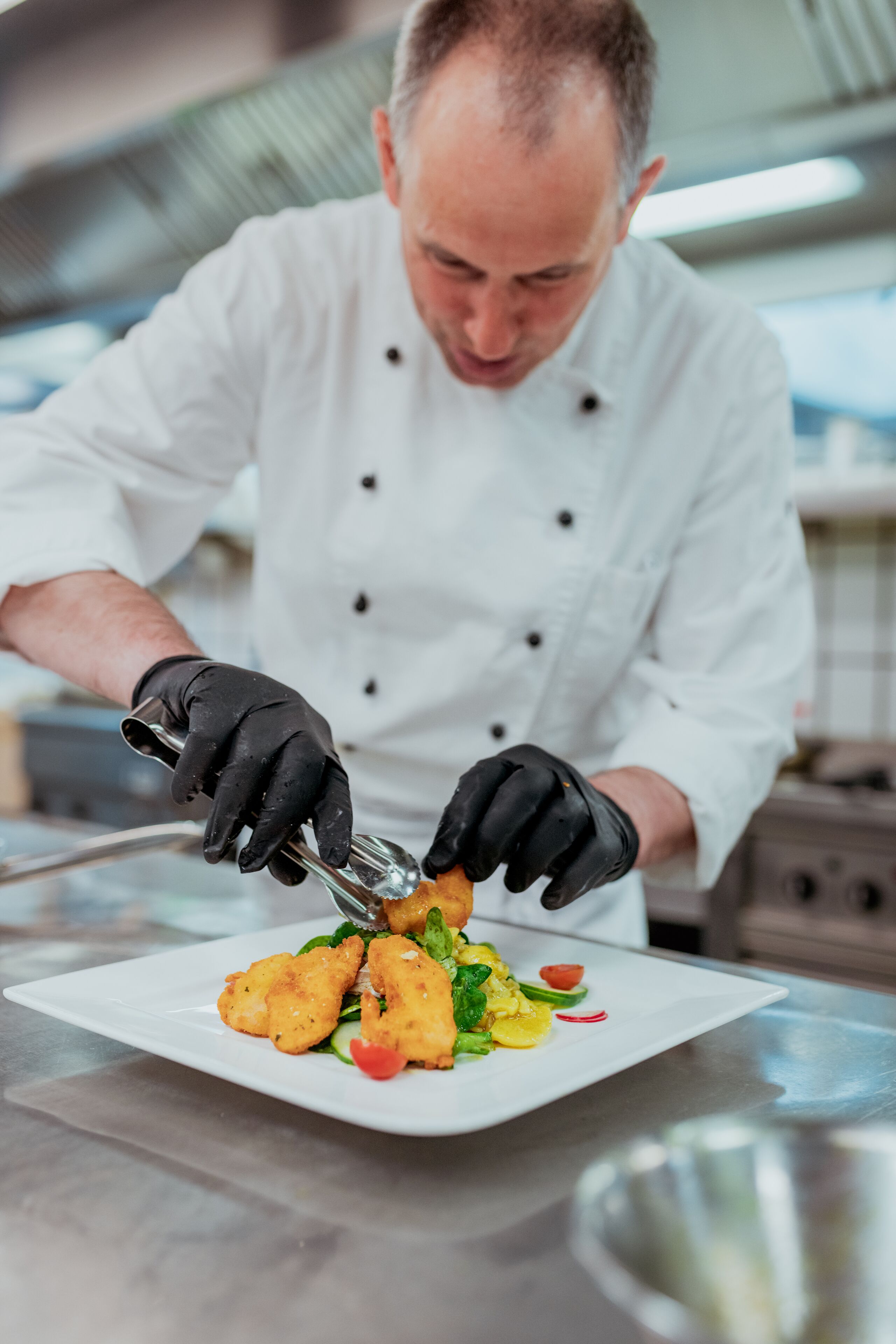 Un chef con uniforme blanco y guantes negros preparando comida en un plato blanco.