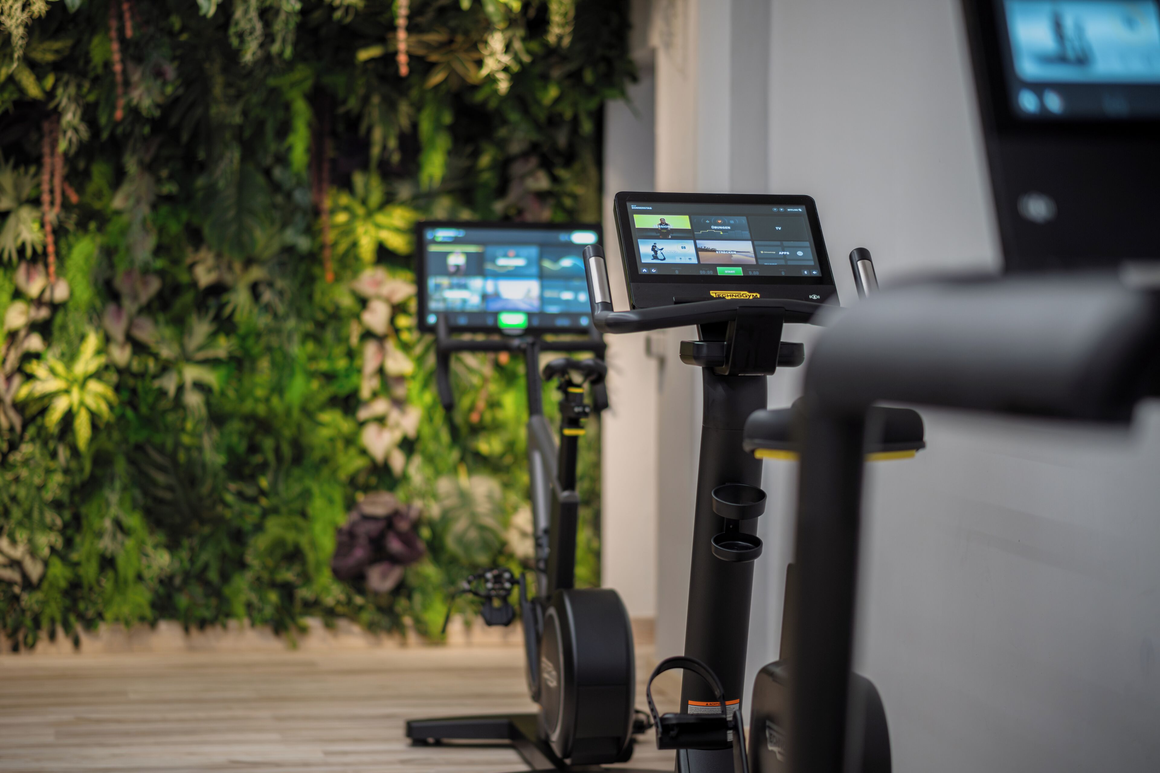 Dos bicicletas estáticas con pantallas digitales, ubicadas en una habitación con una pared cubierta de follaje verde.