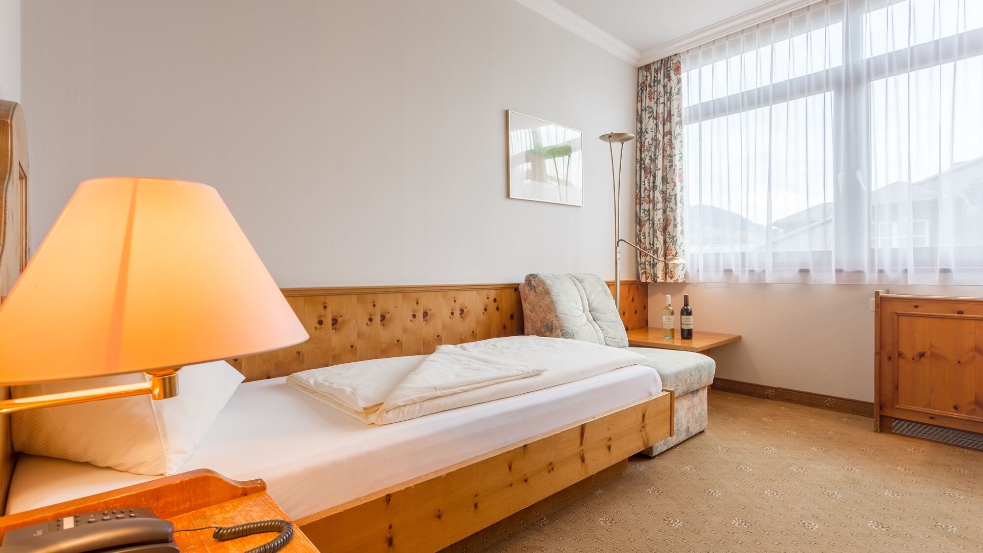 A cozy hotel room with a wooden bed, a bedside table with a lamp, a small table with a bottle, and a window with sheer curtains.