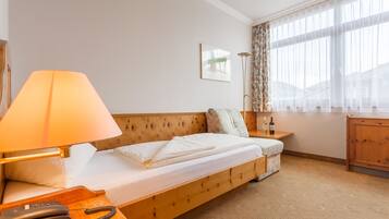 A cozy hotel room with a wooden bed, a bedside table with a lamp, a small table with a bottle, and a window with sheer curtains.
