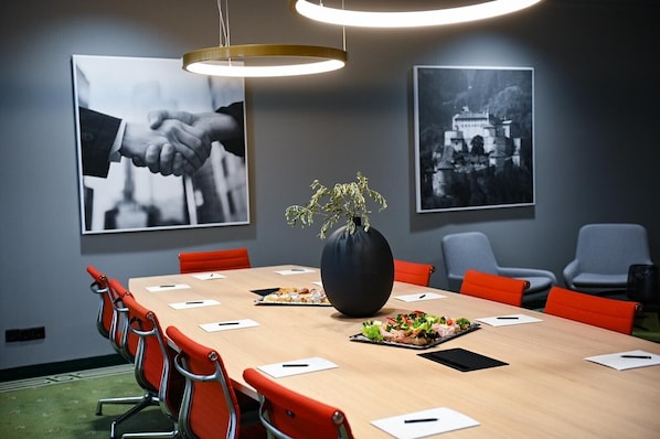 Uma sala de conferências com uma mesa comprida, cadeiras vermelhas, um vaso preto com plantas e duas fotografias preto e branco nas paredes.