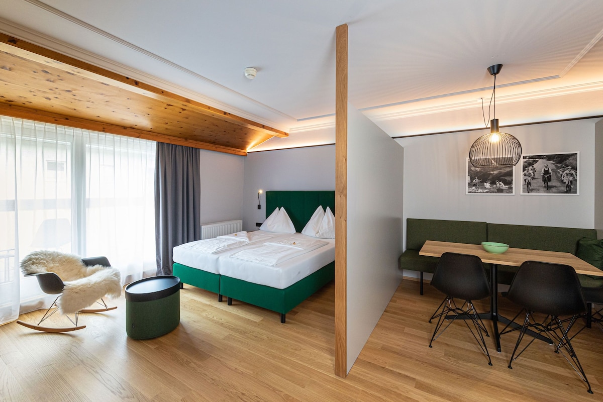 Quarto de hotel moderno com teto de madeira, sofá verde, cama com roupa de cama branca e área de refeições com cadeiras pretas.