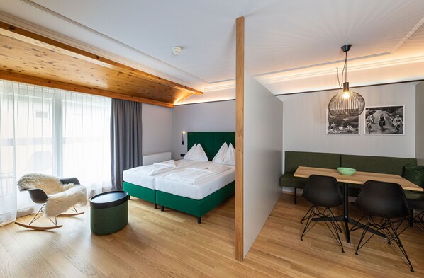 A modern hotel room with a wooden ceiling, a green sofa, a bed with white bedding, and a dining area with black chairs.