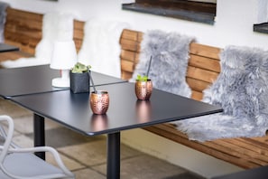 A modern outdoor seating area with a table, two copper mugs with mint leaves, a white lamp, and a wooden bench with a fluffy gray throw.