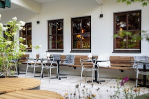 An outdoor seating area with wooden benches and white chairs, a tiled floor, and windows with reflections of trees and lights.