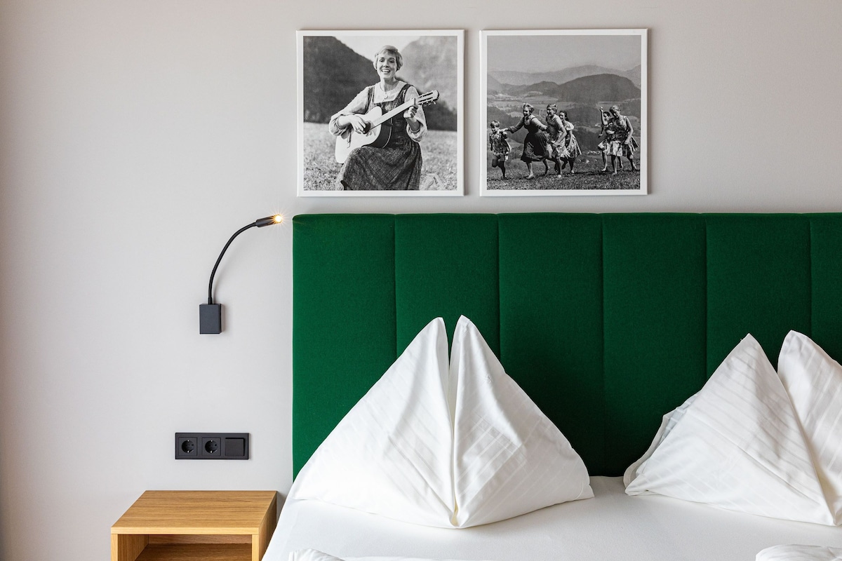 Quarto com cabeceira estofada em verde, almofadas brancas, mesa de cabeceira de madeira e duas fotografias preto e branco na parede.