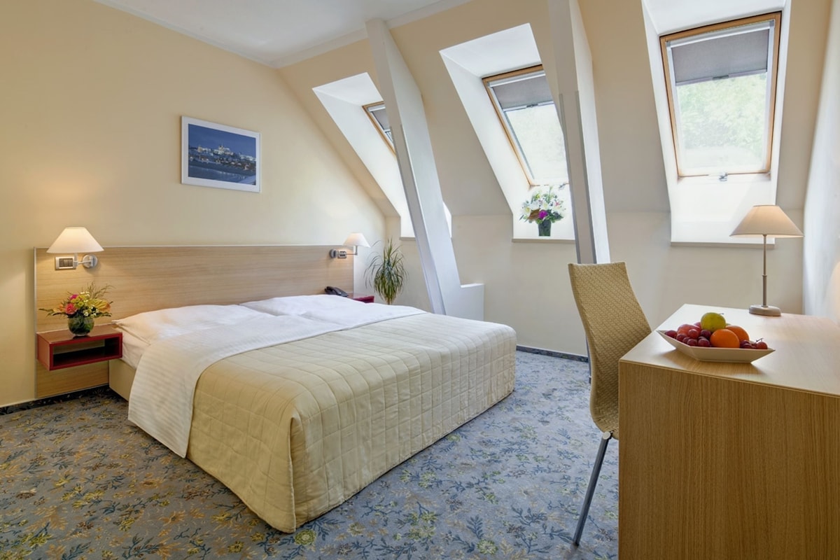 A hotel room with a bed, a desk with a fruit bowl, a chair, a nightstand with a lamp, a picture on the wall, and a window with a view of greenery.