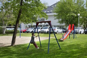 A playground with swings and slides, surrounded by trees and parked cars. - Hotel Michael (Prague)