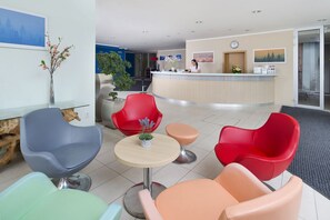 A modern hotel lobby with a reception desk, red and blue chairs, a small round table, and a vase with flowers. - Hotel Michael (Prague)