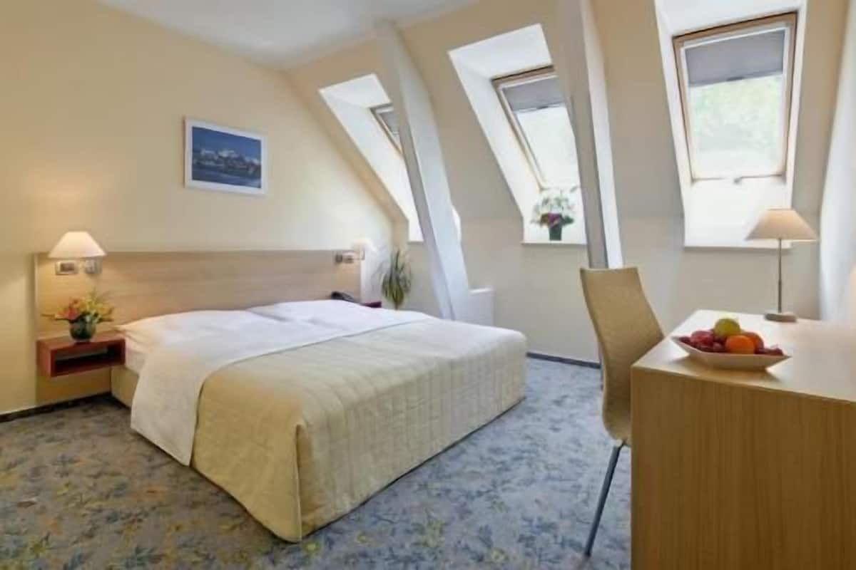 Hotellihuone, jossa on sänky, työpöytä, jossa on hedelmäkulho, tuoli, yöpöytä lampun kanssa ja ikkuna, josta on näkymä.