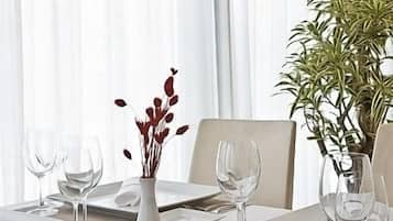 Mesa de comedor preparada para cuatro personas, con platos blancos, cubiertos y copas de vino. Hay un florero con flores rojas y una planta en maceta sobre la mesa.