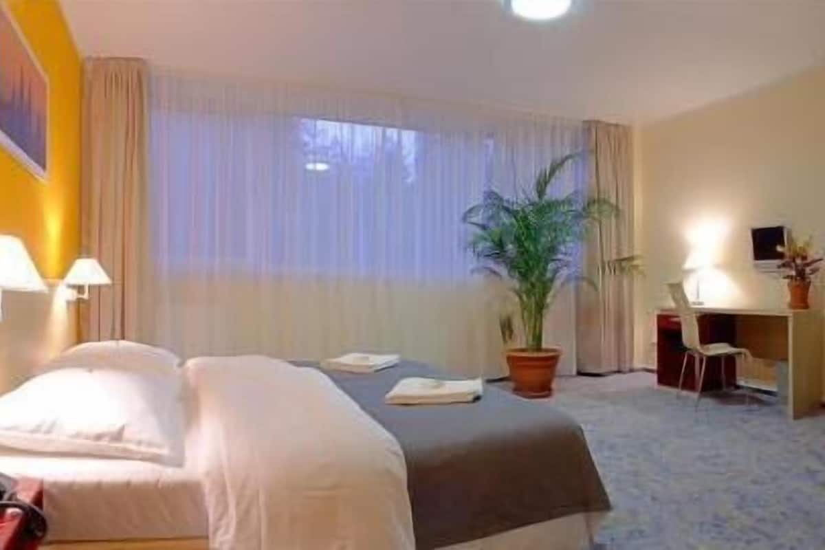 Hotellihuone, jossa on sänky, yöpöytävalaisimet, työpöytä tuolilla, ruukkukasvi ja ikkuna, jossa on verhot.
