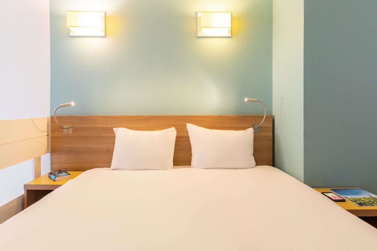 A hotel room with a wooden headboard, two bedside lamps, a bed with white linens, and a nightstand with a book and a smartphone.