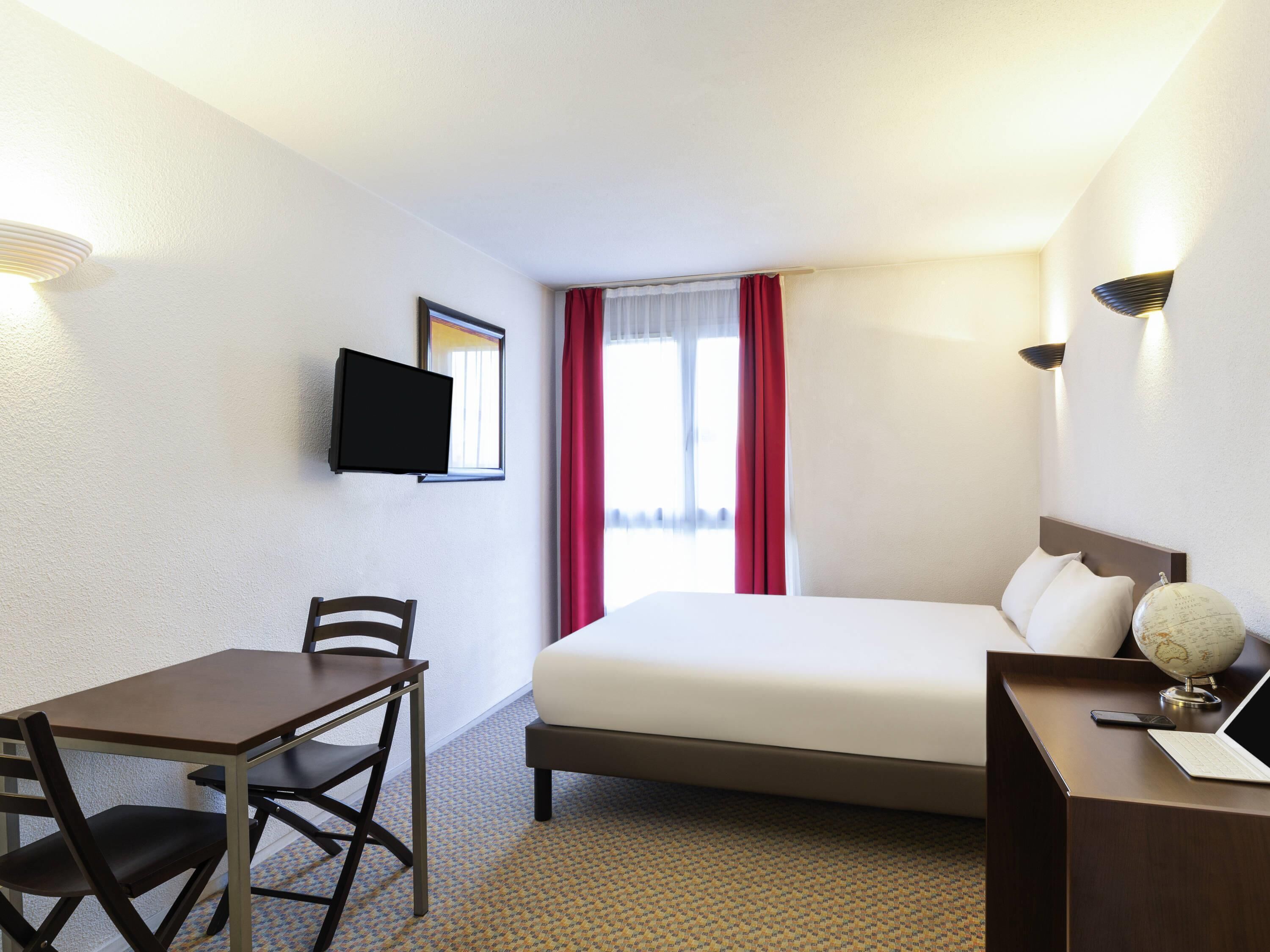 A hotel room with a bed, a desk with a chair, a television, and a globe on the nightstand.