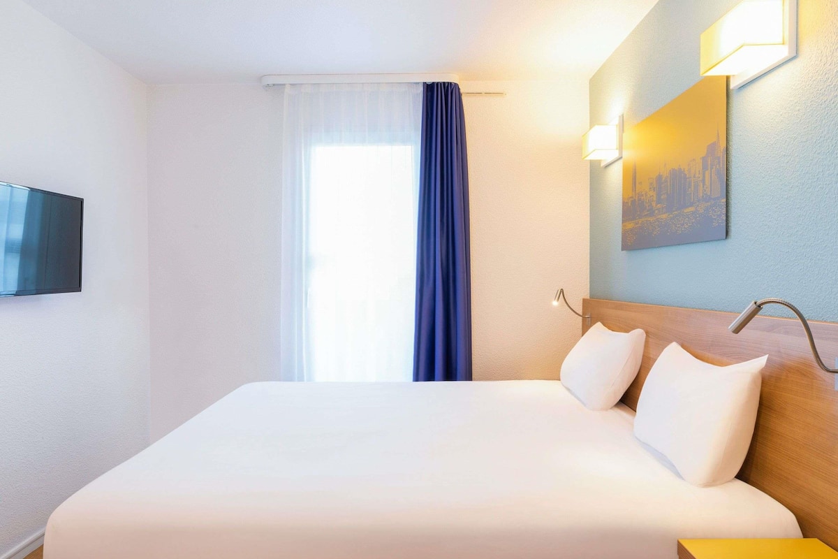 A hotel room with a bed, a television, a painting, and a window with curtains.