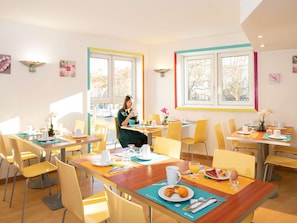 Uma sala de jantar luminosa com várias mesas, cadeiras e uma pessoa sentada à uma delas.