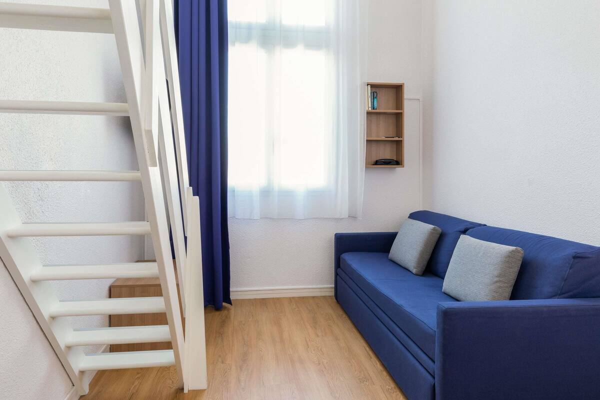 A room with a white staircase, a blue sofa, a wooden shelf, and a window with curtains.