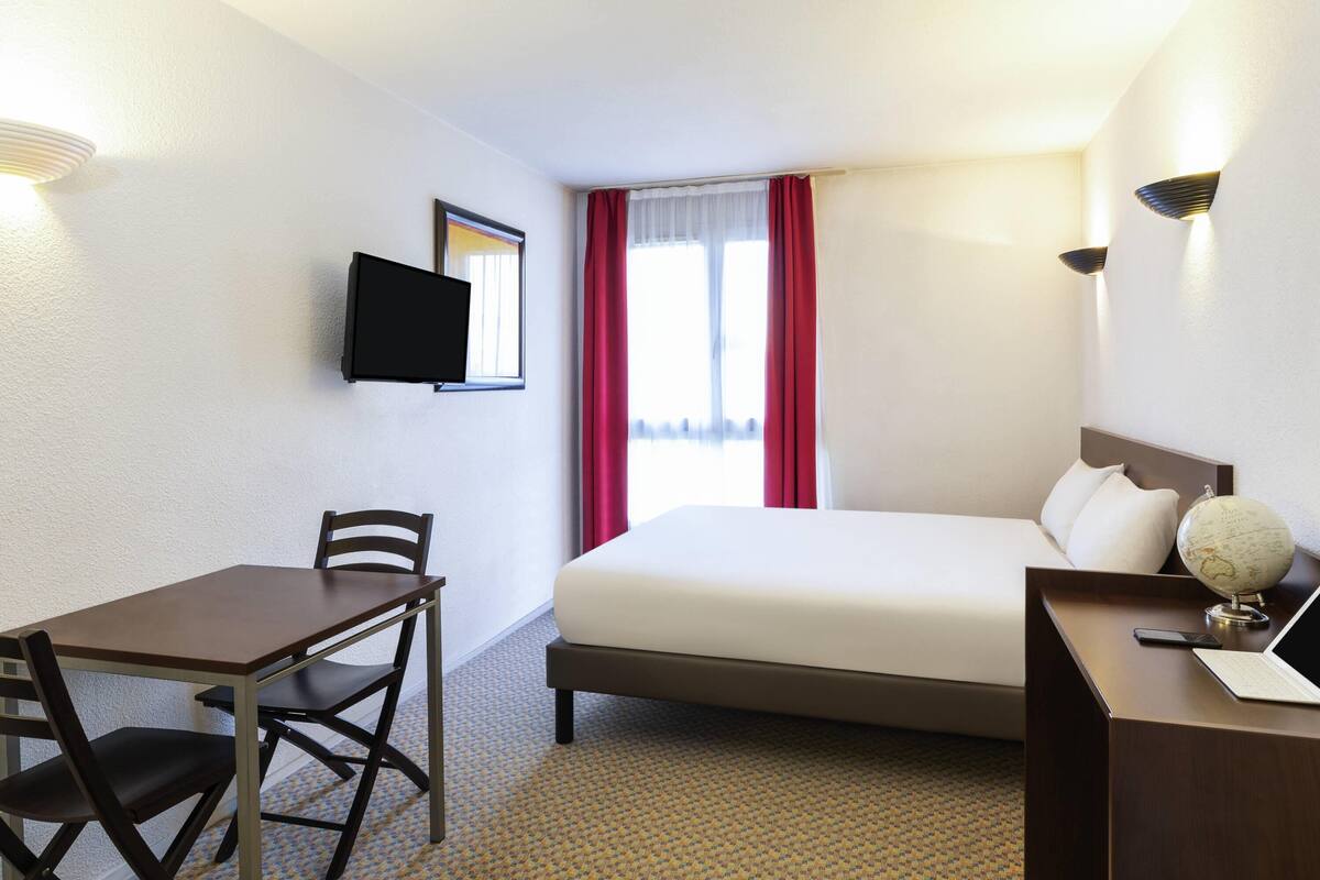 A hotel room with a bed, a desk with a chair, a television, and a globe on the nightstand.