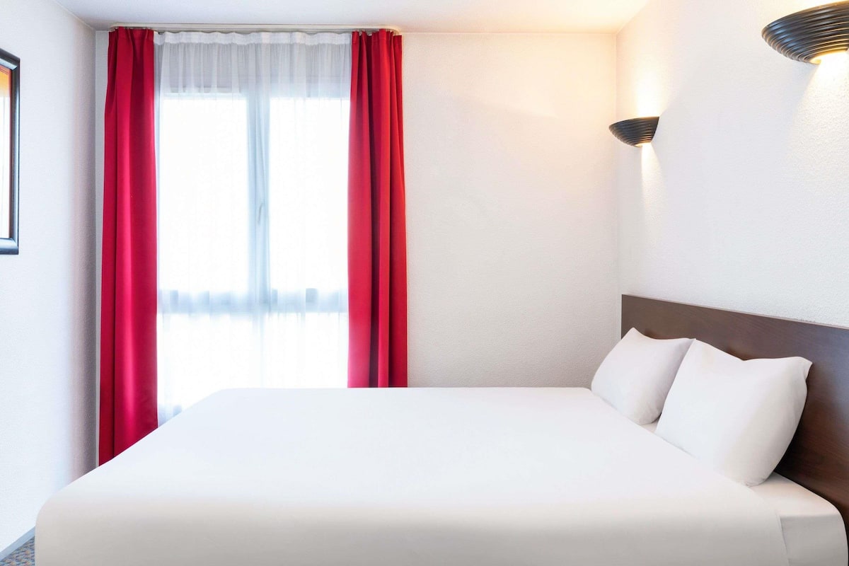 A hotel room with a large bed, red curtains, a wall-mounted light, and a framed picture on the wall.