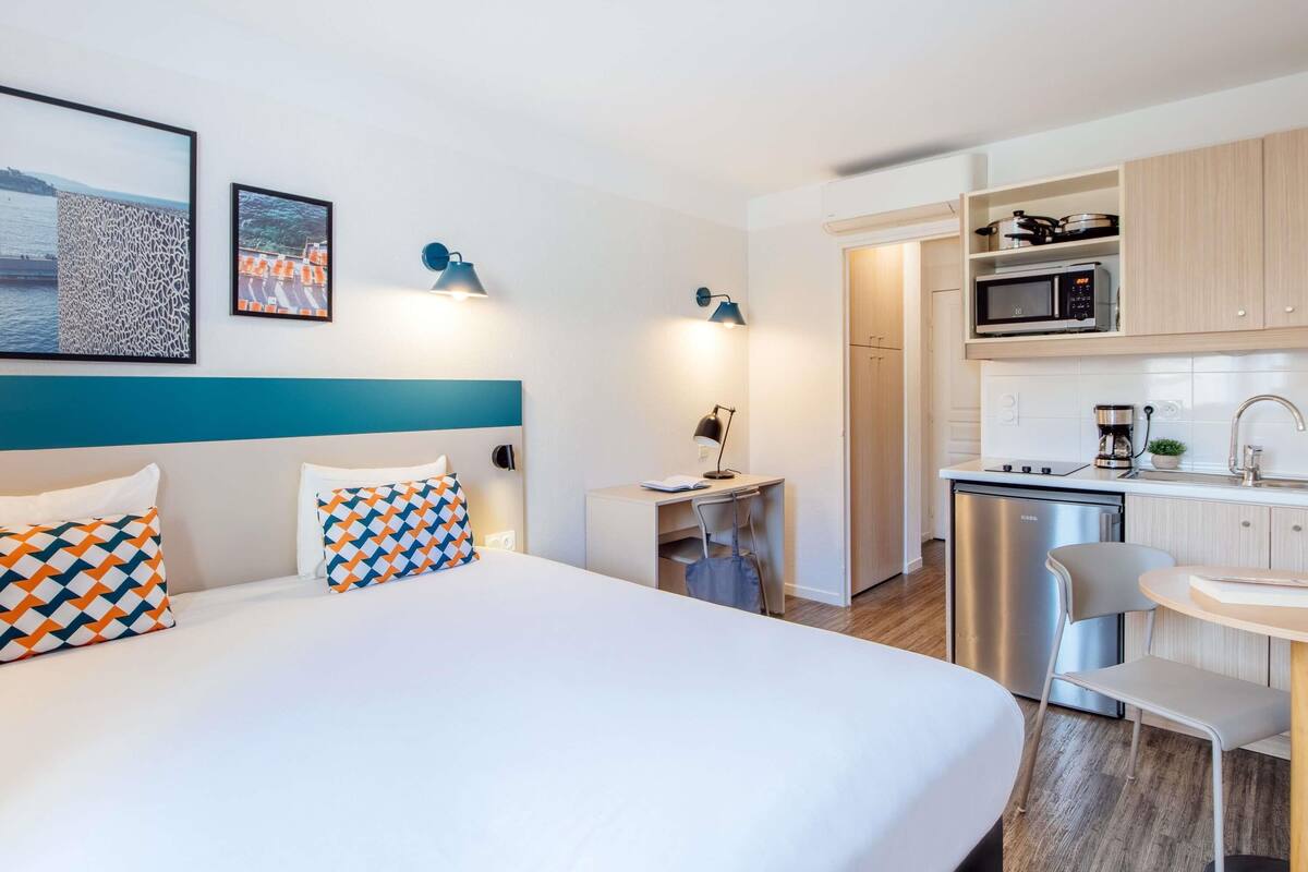 Une chambre d’hôtel comprenant un lit, un bureau, une chaise, un petit coin cuisine avec un four à micro-ondes et une machine à café, ainsi qu’une porte donnant sur une autre pièce.