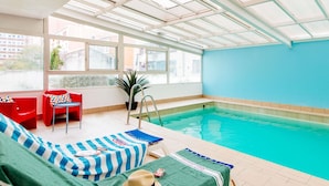 Ein Innenpool mit Glasdach, roten und grünen Liegestühlen und einem blauen Tisch am Beckenrand.