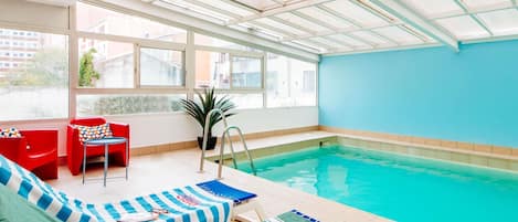 Ein Innenpool mit Glasdach, roten und grünen Liegestühlen und einem blauen Tisch am Beckenrand.