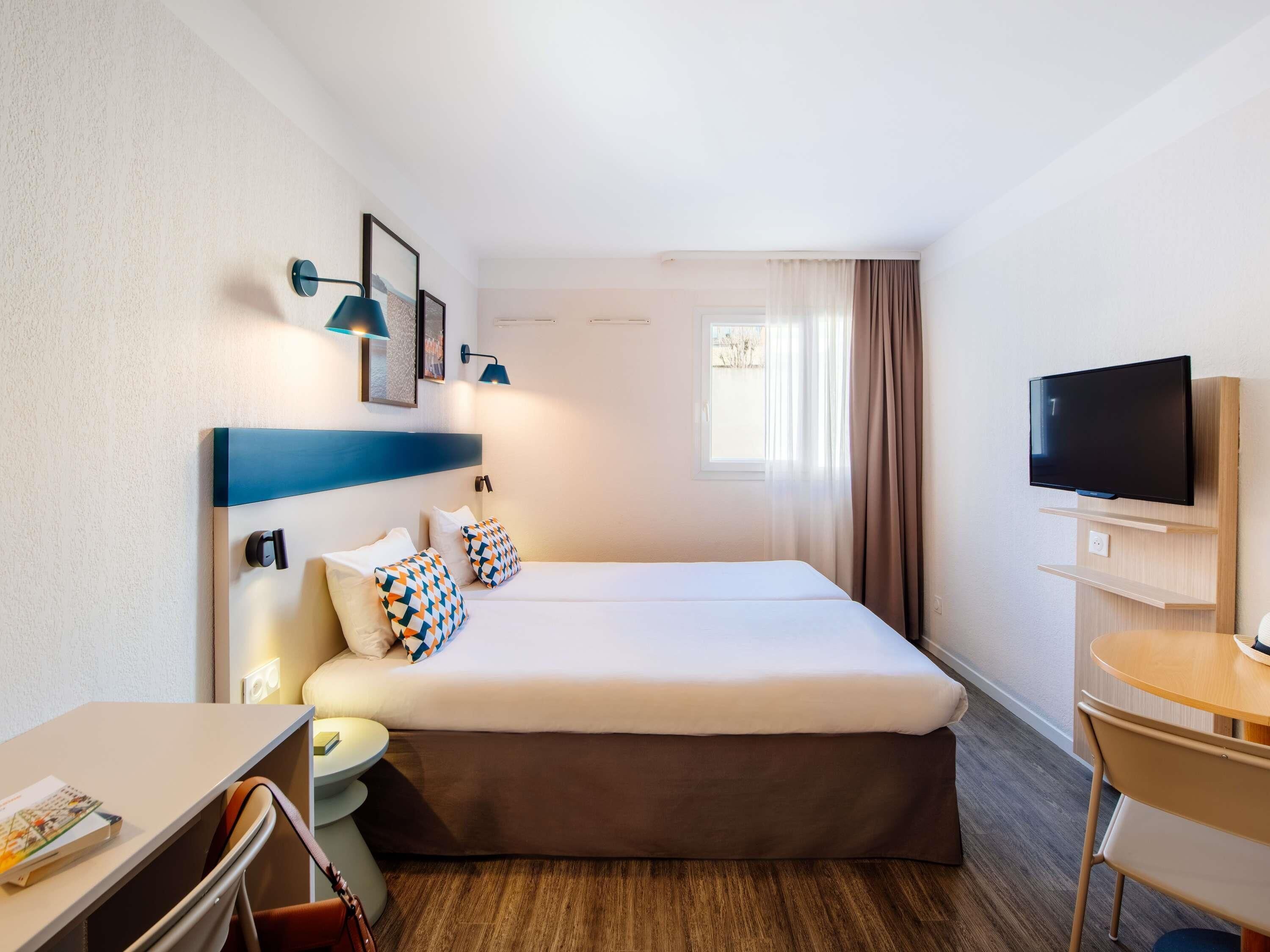 A hotel room with a bed, bedside table, lamp, TV, and a window with curtains.