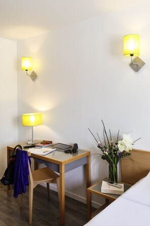 A hotel room with a wooden desk, two wall-mounted lamps, a vase with white flowers, a chair with a blue jacket, and a bedside table with a book and remote controls. - Aparthotel Adagio Access Marseille Prado Perier (Marseille)