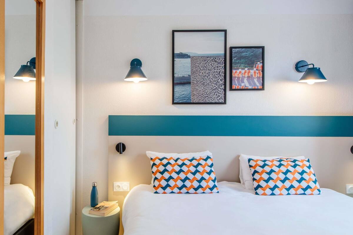 Une chambre d’hôtel avec un lit, deux lampes fixées au mur, un tableau encadré, une table de chevet avec une bouteille et une petite table d’appoint.