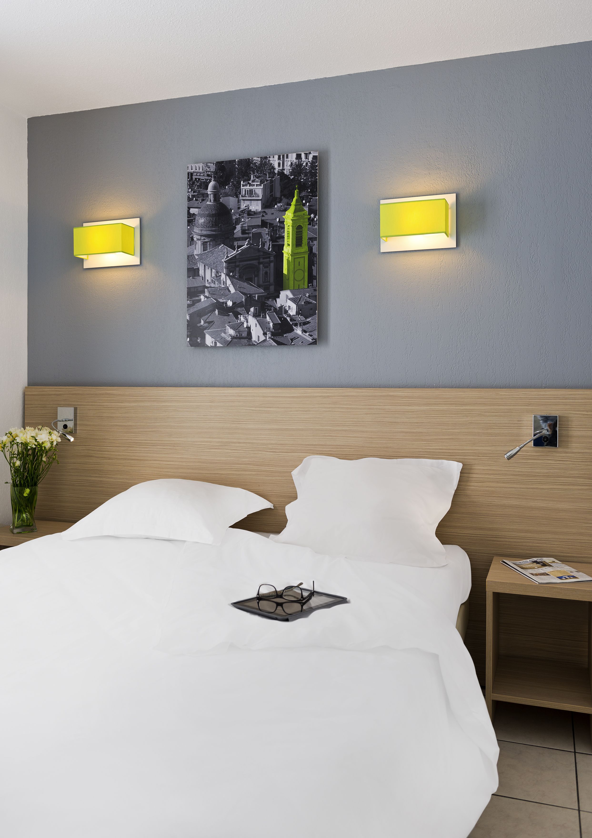 Una habitación de hotel con una cama, dos lámparas de pared, un cuadro con una vista urbana enmarcada y una mesita de noche con una revista.
