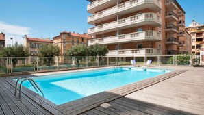 Une piscine dotée d’une terrasse en bois, entourée d’une clôture, et située à côté d’un immeuble à plusieurs étages avec des balcons.
