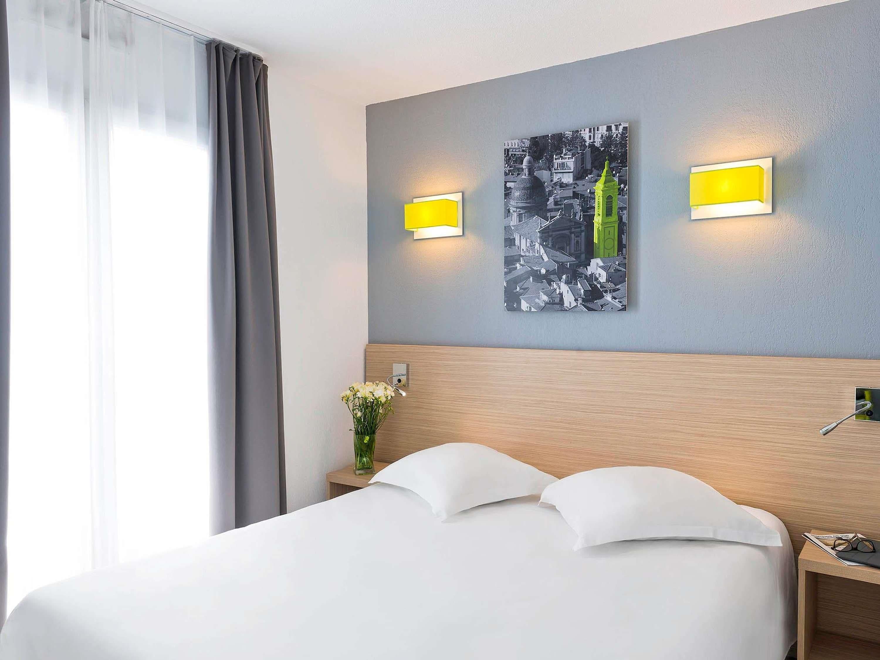 A hotel room with a bed, two bedside lamps, a nightstand, a vase with flowers, and a wall-mounted picture.