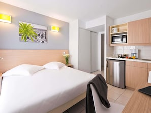 Hotelkamer met een bed, nachtkastje, wandlamp, schilderij en een kitchenette met magnetron, koffiezetapparaat en spoelbak.