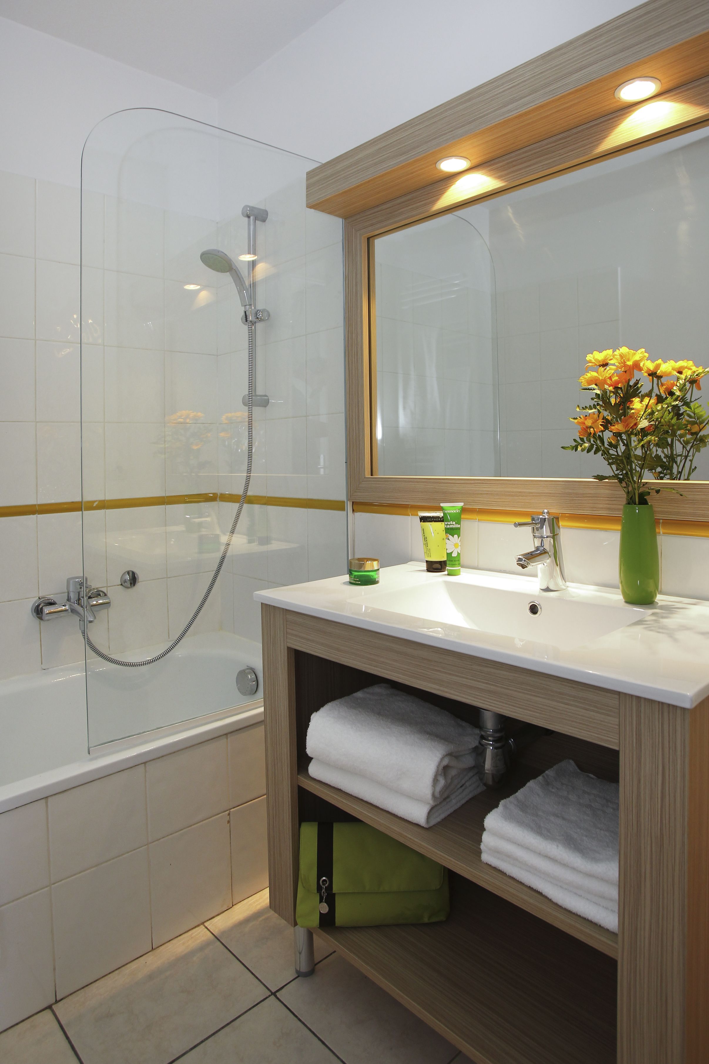 A bathroom with a glass shower enclosure, a white sink with a mirror above it, and a green vase with flowers.