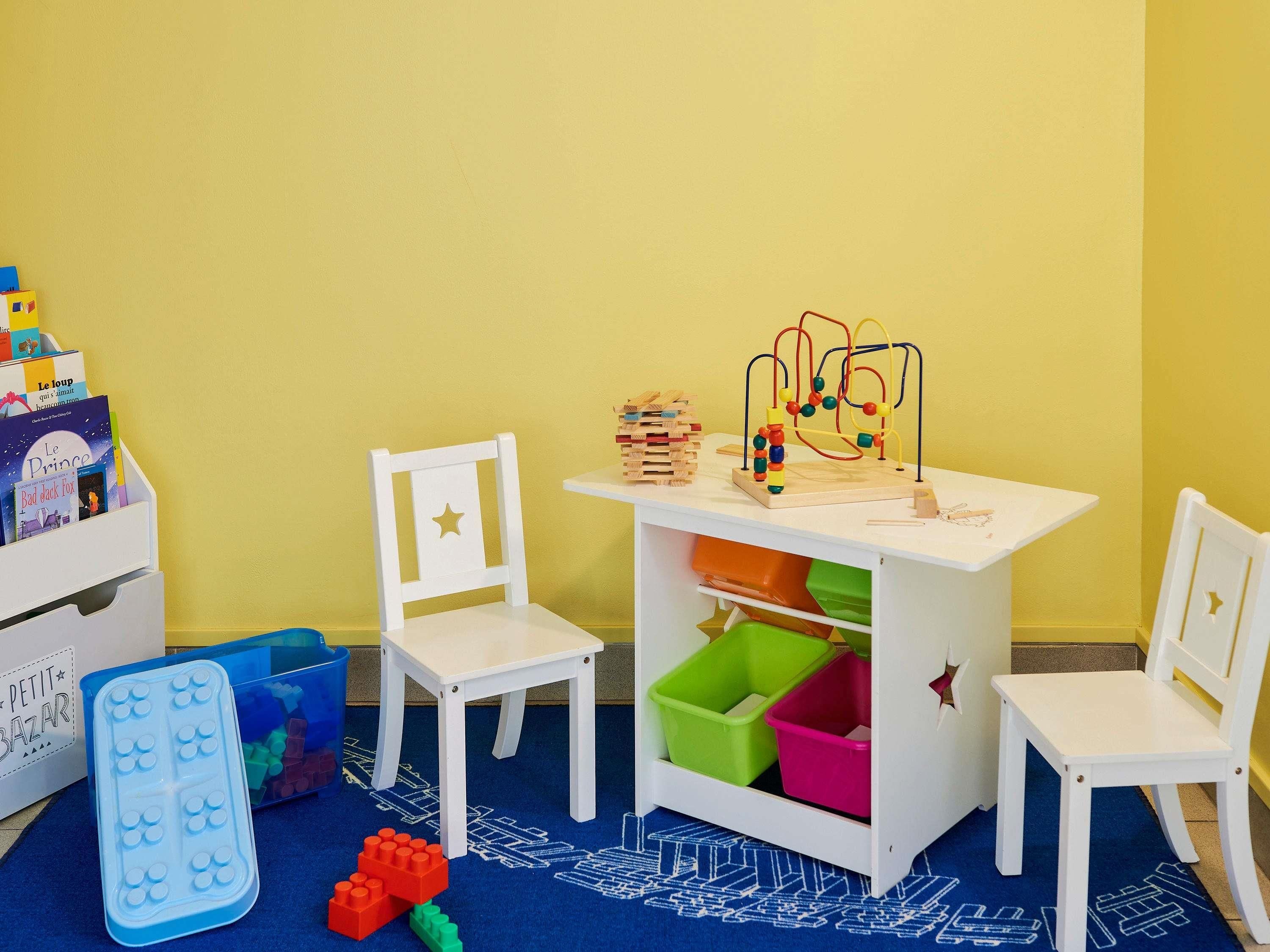 Ein Spielbereich für Kinder mit zwei weißen Stühlen, einem kleinen Tisch mit Spielzeug und Aufbewahrungsboxen.