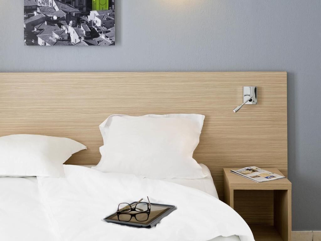 A neatly made bed with white bedding, a bedside table with a lamp, and a wall-mounted power outlet.